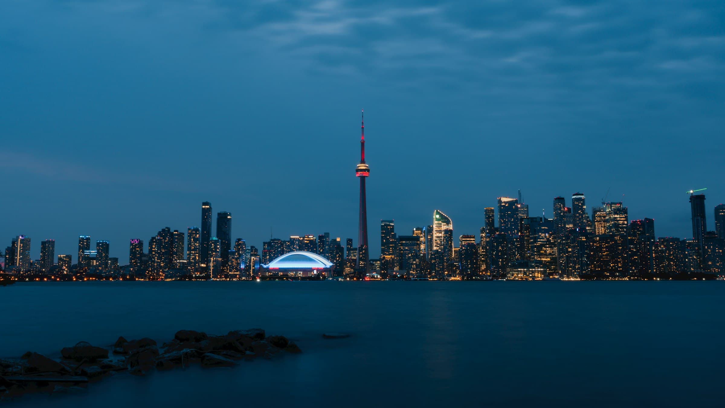 The Toronto skyline lit up at blue hour, with the CN Tower in red, seen across Lake Ontario